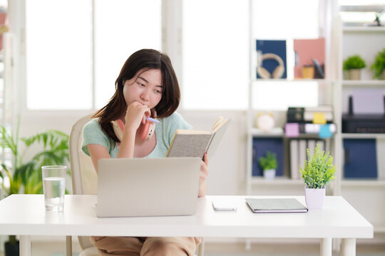 Asian adult woman studies reads book research on laptop computer remote learning at home office desk education online training