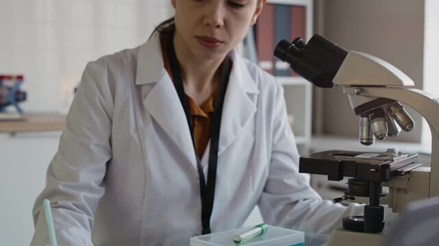Tilt up shot of female young laboratory technician in white coat filling out form while analyzing sample in microscope at table