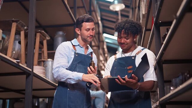 Two diverse male colleagues discussing and checking stock list using clipboard and barcode scanner in warehouse. Concept of teamwork, logistics, and inventory management.