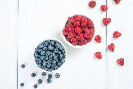 bilberry and raspberry fruits at white ceramic cup, on bright wood table