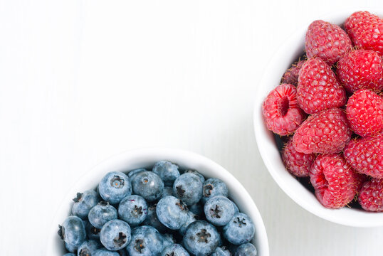 bilberry and raspberry fruits at white ceramic cup, on bright wood table
