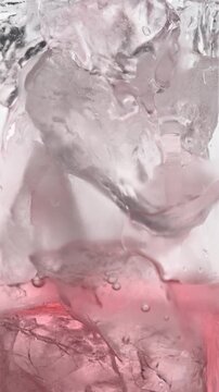Macro view of pink sparkling drink flowing through clear ice cubes in glass, refreshing beverage background in vertical frame