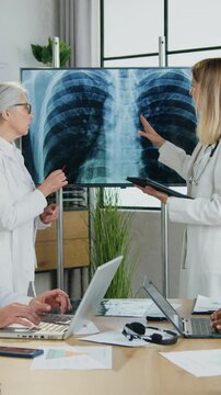 Beautiful respected senior successful female head doctor talking with younger female colleague about chest and lungs radiography while qualified smart male workmates working with computers