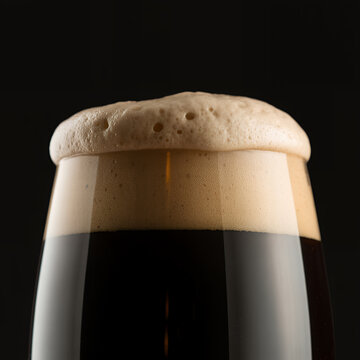 Close up of a glass filled with dark beer and a thick layer of foam against a dark background