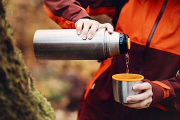 Naklejka premium Hands pour hot liquid from thermos into cup with smiling woman in background. tea break outdoors, coffee on hike, hot drink in forest, warming up outdoors.