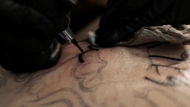 Close-up of a female tattoo artist in black latex gloves using a wireless rotary tattoo pen to ink a large, intricate floral and mandala design on a woman's upper back.