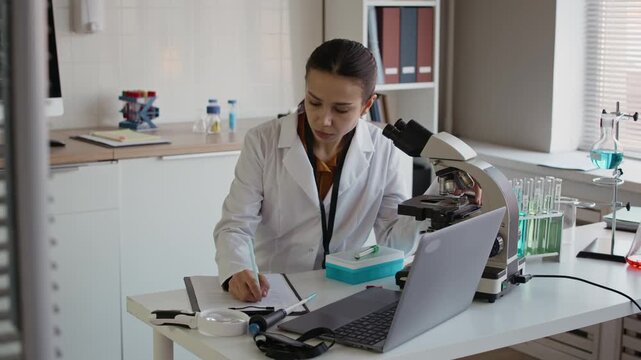 Medium shot of young Caucasian female scientist in white coat using microscope and taking notes while sitting at table with wireless laptop