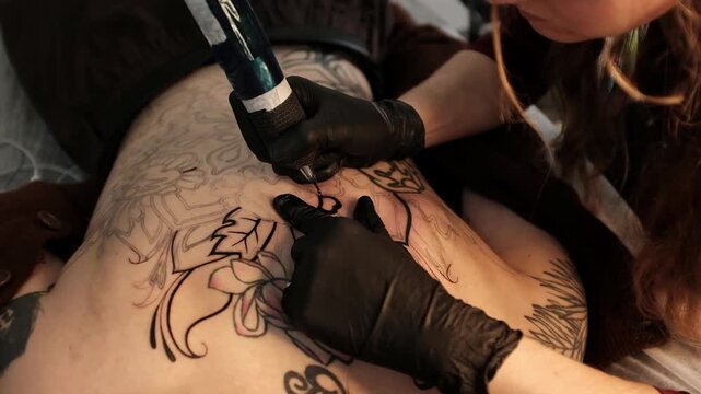 Close-up of a female tattoo artist in black latex gloves using a wireless rotary tattoo pen to ink a large, intricate floral and mandala design on a woman's upper back.