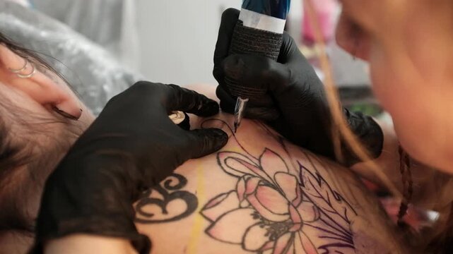 Close-up of a female tattoo artist in black latex gloves using a wireless rotary tattoo pen to ink a large, intricate floral and mandala design on a woman's upper back.