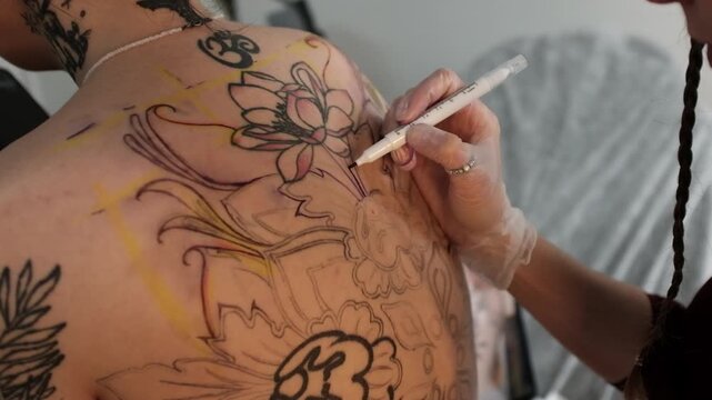 Close-up of a female tattoo artist draws a scketch of intricate floral and mandala design on a woman's upper back. Free hand tattoo technique.