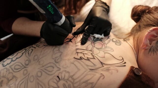 Close-up of a female tattoo artist in black latex gloves using a wireless rotary tattoo pen to ink a large, intricate floral and mandala design on a woman's upper back.