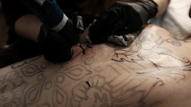 Close-up of a female tattoo artist in black latex gloves using a wireless rotary tattoo pen to ink a large, intricate floral and mandala design on a woman's upper back.