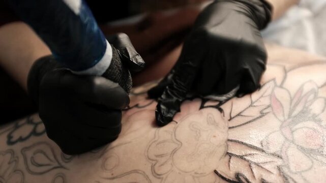 Close-up of a female tattoo artist in black latex gloves using a wireless rotary tattoo pen to ink a large, intricate floral and mandala design on a woman's upper back.