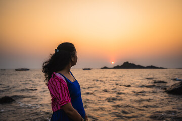 Fototapeta premium Silhouette of a woman standing by the sea during sunset with waves and rocky shoreline