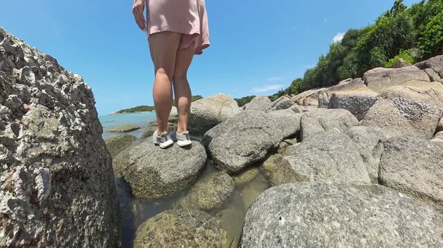 Woman stepping across wet rocks by shoreline with sea visible under bright sunlight camera low angle Adventure and Coastal Trekking Concept