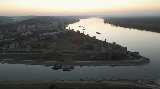 Drone retreats high above Smederevo Fortress revealing its dark silhouette against a warm dusk sky over the Danube river, Smederevo, Serbia.