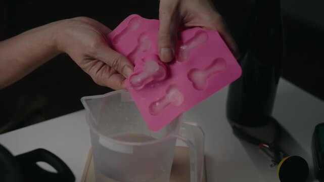 Close-up view of unrecognizable person hands pushing out frozen ice cubes from pink silicone tray into pitcher filled with water, preparing refreshing drink or treat. Shooting in slow motion.