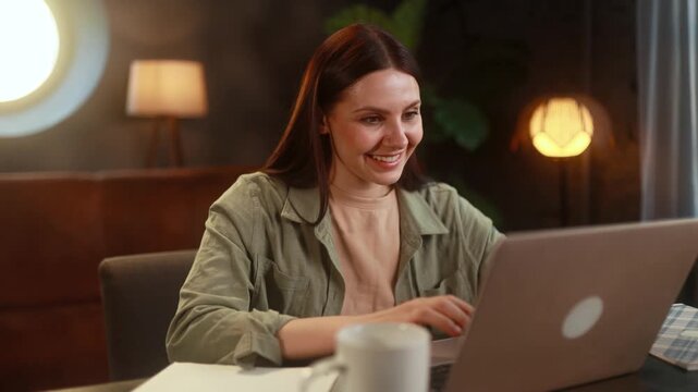 Smiling young woman working online on her laptop, looking happy and engaged while completing tasks at home in a comfortable indoor environment