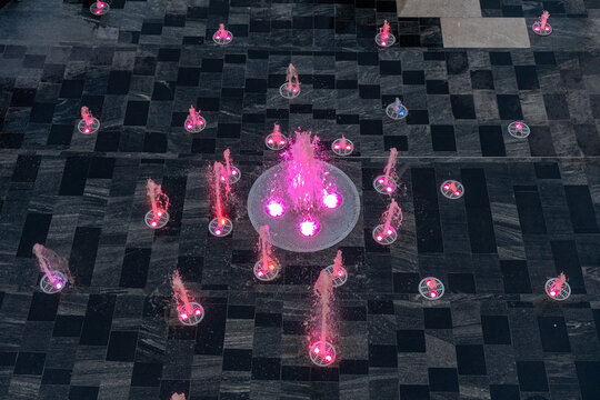 Top view of a modern fountain with pink LED lights embedded in a tiled floor at an outdoor shopping mall, creating a geometric and decorative pattern