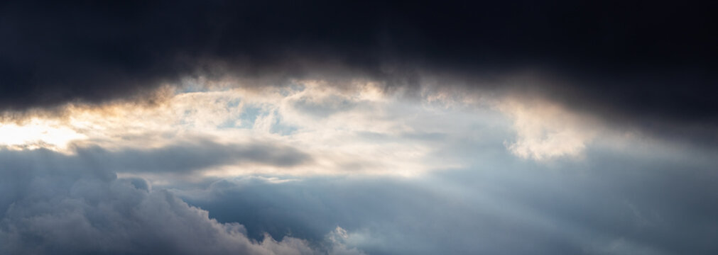 Dramatic stormy sky with dark clouds and rays of light through the gap, panoramic view of thunderclouds