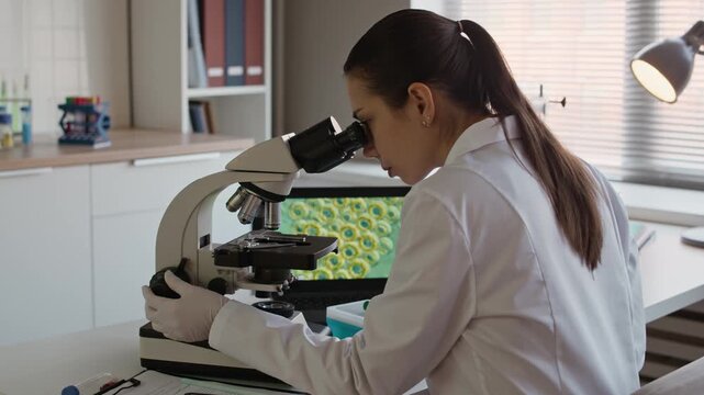 Medium shot of young female Caucasian microbiologist using microscope while examining sample at table with cell structure on computer screen in modern laboratory
