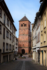 Obraz premium Historic street Saverne Alsace toward red Notre-Dame-de-la-Nativite church tower clock cross. Traditional European architecture cobblestone road colorful facades calm urban atmosphere dramatic sky