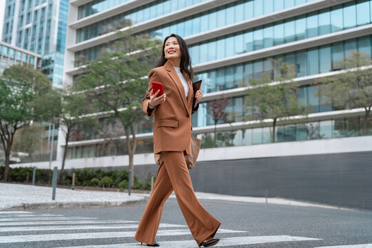 Asian businesswoman using smartphone on city street, professional career woman checking online banking and finance apps, walking near business office buildings in urban downtown. Full body, copy space