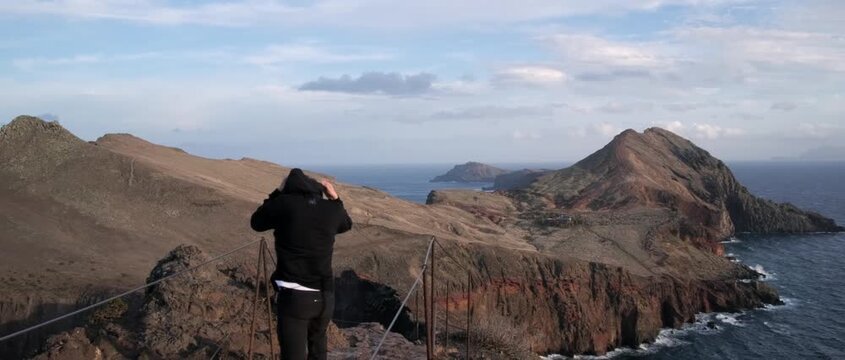 View of a mountain range overlooking the ocean, with two people walking along a trail under a clear sky. Scenic landscape of Madeira, Portugal, at Ponta de S&atilde;o Louren&ccedil;