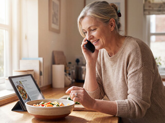 smiling middle aged woman talking on smartphone while cooking healthy meal in kitchen as concept of digital lifestyle multitasking smart home technology and modern hobby with online recipe on tablet