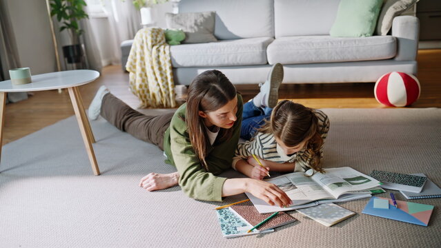 Mother assisting daughter studying school subject lying apartment floor together