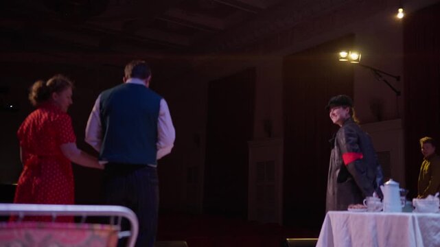 Theatrical confrontation on dimly lit stage as costumed performers gather near elegant tea table during dramatic play rehearsal in vintage styled auditorium