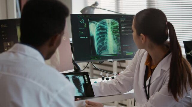 Medium shot of biethnic young colleagues in white coats discussing chest CT on screens of digital devices in modern clinic office