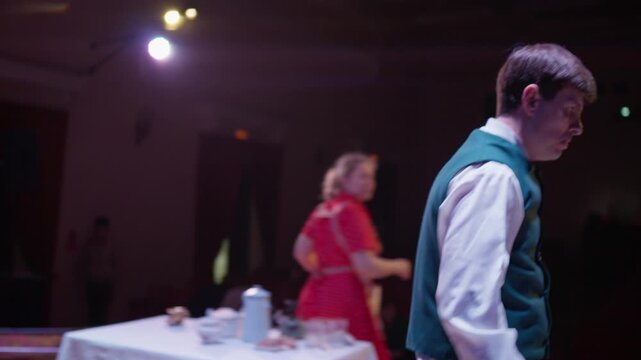 Amateur stage performance with actors in vintage costumes around a white table set for tea in a dimly lit theater with an empty audience and dramatic lighting