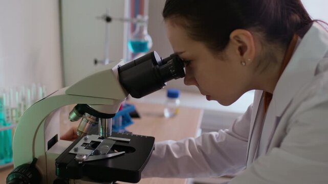 Handheld shot of young Caucasian technician examining molecules in microscope connected to computer monitor with real time image in modern laboratory