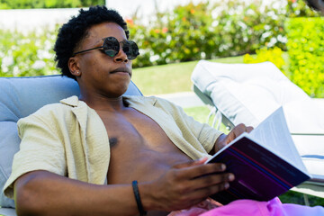 Fototapeta premium African American man reclining, reading book on pool chair, wearing sunglasses and open beige shirt