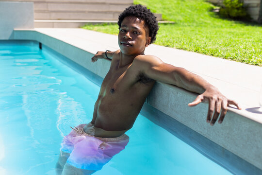 African American man leaning on coping wearing pink trunks, gazing in clear-blue yard pool by steps