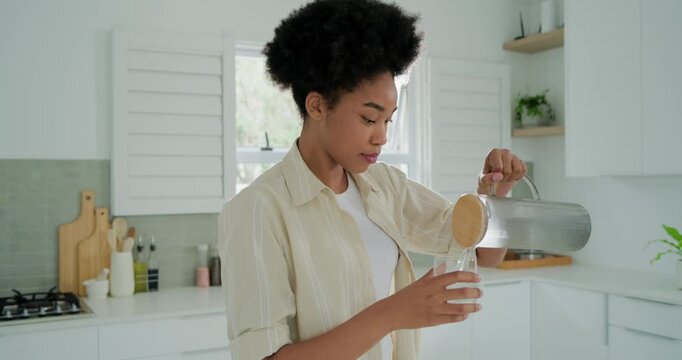 Adult African woman lifting pitcher pouring into glass, hydrating and checking phone in kitchen