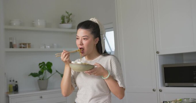 African American woman eating bowl of noodles with chopsticks in home kitchen, hunger driving bites