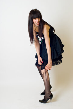Studio portrait of a young woman wearing an elegant black dress, posing against a clean white background. The model is captured in a full-length or three-quarter pose, exuding confidence