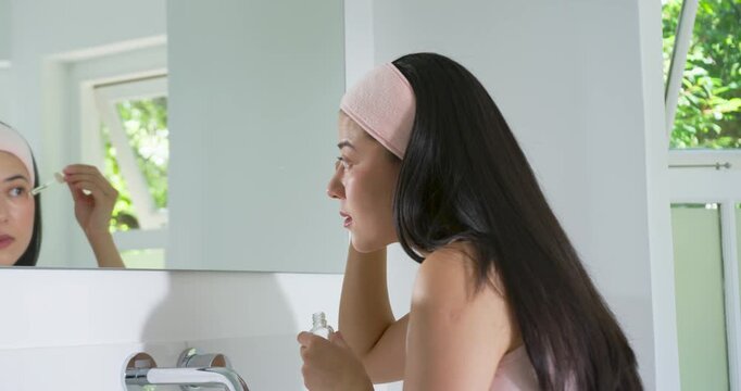 Woman leaning at vanity checking mirror before using dropper applying skincare serum, copy space
