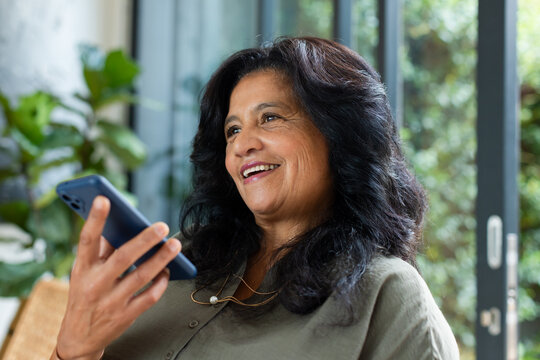 African American mature woman smiling and holding smartphone near face in sunroom with plants