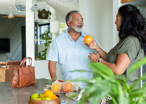 Couple standing at stone countertop in kitchen handing orange near leather handbag and mesh bags