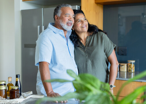 Senior couple standing in kitchen, leaning, smiling, wearing light blue and olive, fridge and watch