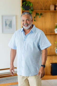 Senior African American man standing smiling at home wearing light blue shirt, wooden shelf plants