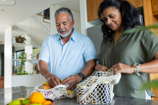 African American senior couple sorting and placing citrus into white mesh bags at kitchen island