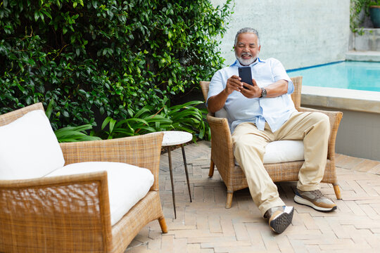 Senior African American man smiling, holding smartphone on brick poolside patio by pool, copy space