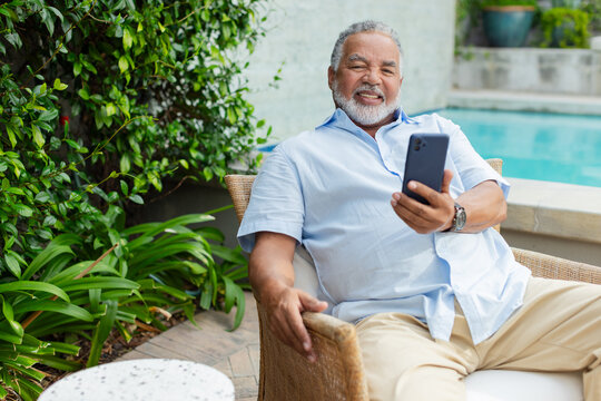 Senior African American man relaxing in wicker chair on pool deck, holding phone and wearing watch