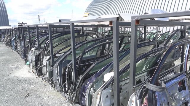 Rows of car doors on car disassembly racks. Metal frames, different colors, daylight, industrial site.