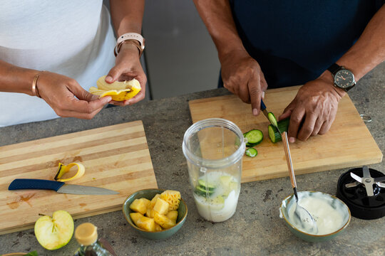 African couple peeling banana and slicing cucumber at home on wooden boards, blender cup