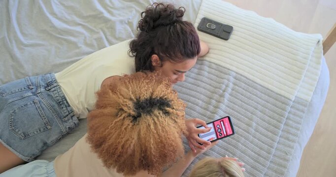 Vertical video: Tapping phone, three friends scrolling pink shopping app on bed, selecting outfits